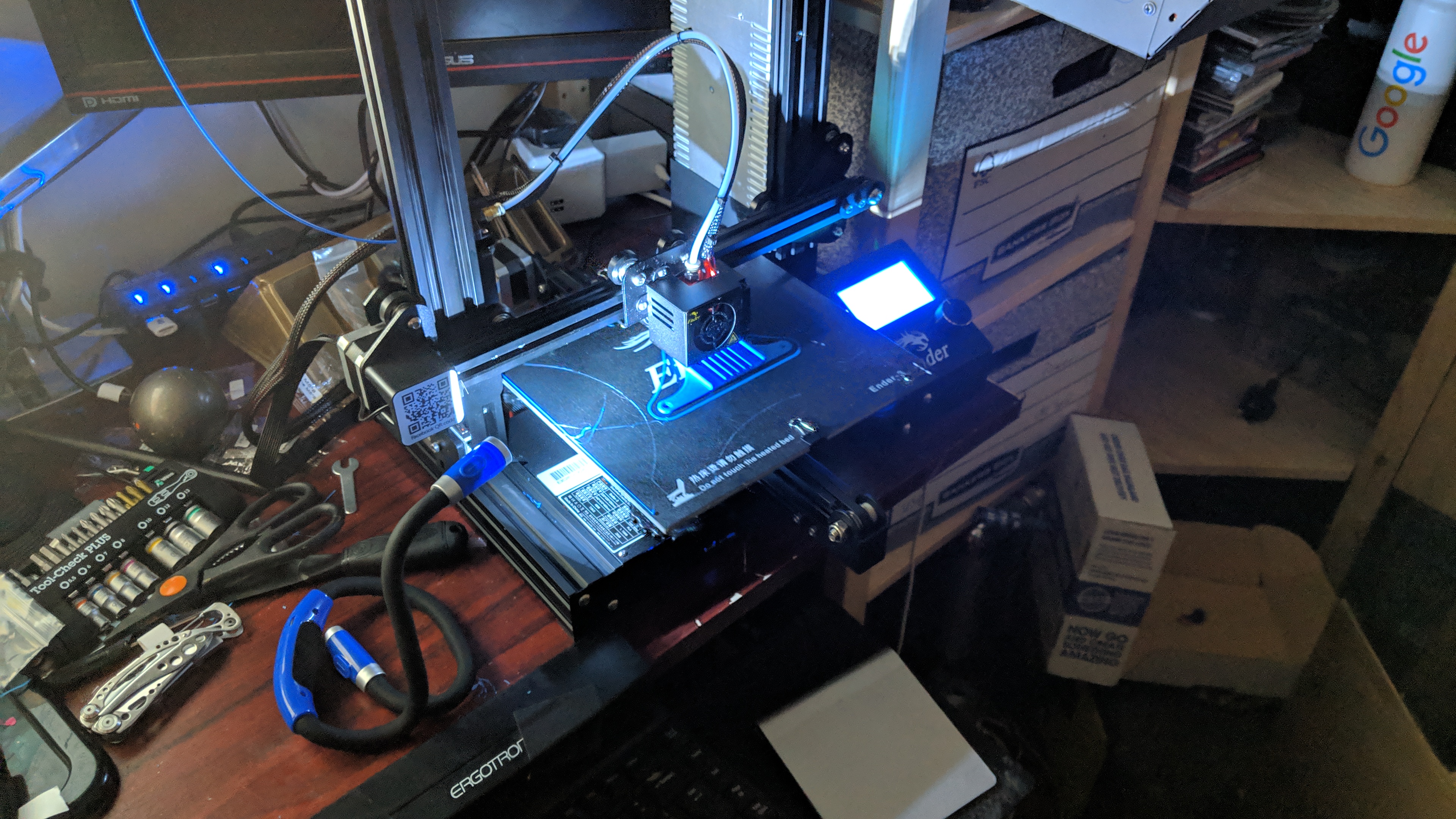 A black 3D printer on a desk, part-way through a fan shroud print in the same blue plastic as the previous image, but looking much neater